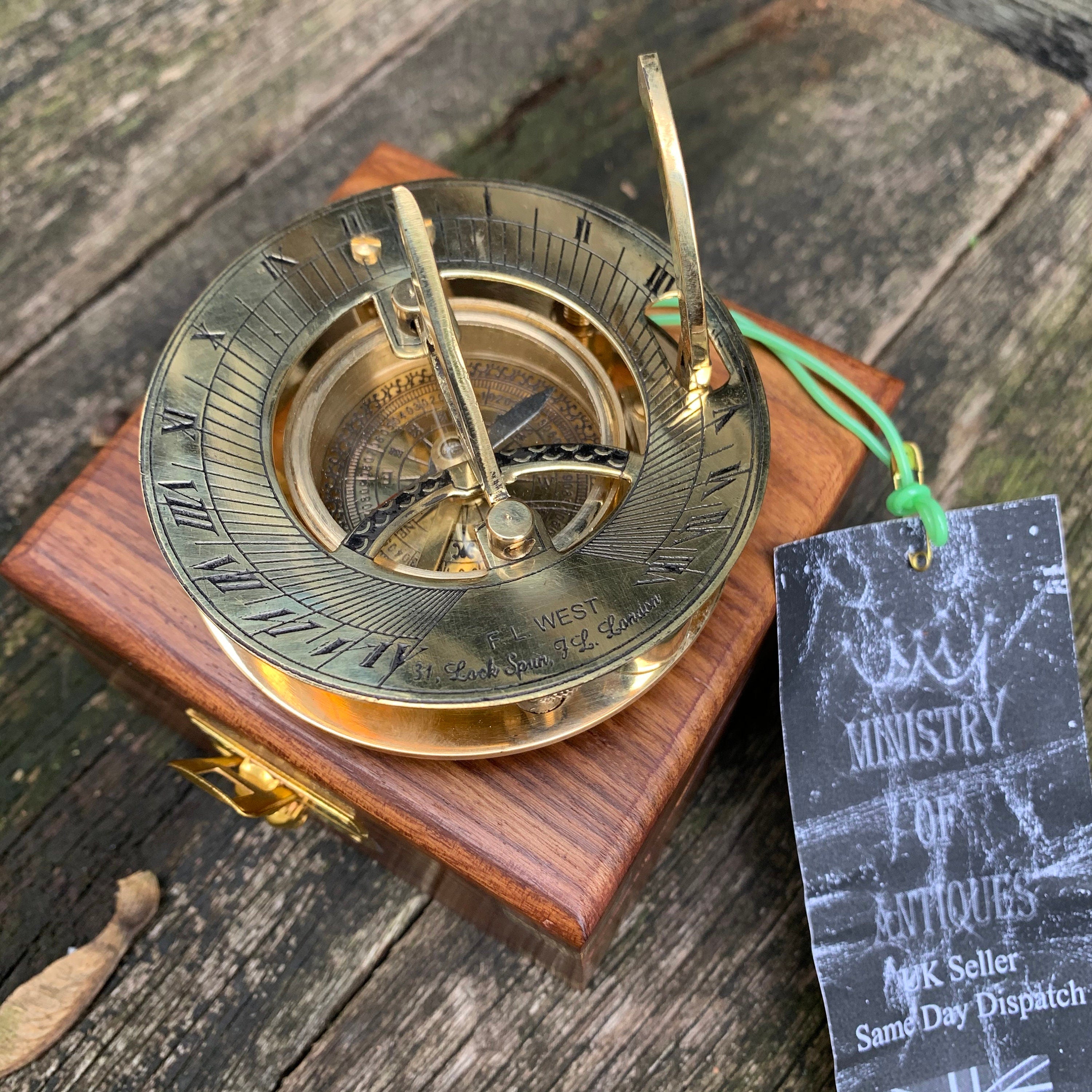 Puede incluir: Un reloj de sol de latón antiguo con números romanos y una brújula, montado sobre una base de madera. El reloj de sol tiene un gnomon y texto grabado. Una etiqueta dice "MINISTRY OF ANTIQUES".