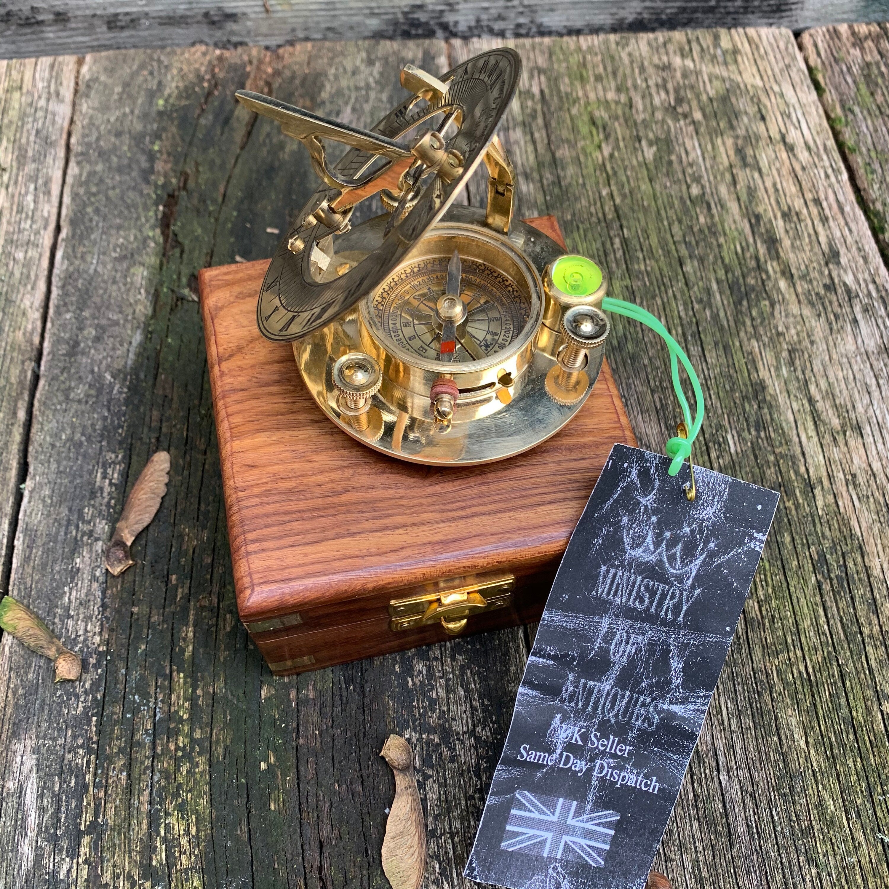 Puede incluir: Una brújula de reloj de sol de latón sobre una caja de madera. La brújula presenta una esfera detallada y un gnomon. Se adjunta una etiqueta con el texto "MINISTRY OF ANTIQUES". Es un instrumento de navegación.