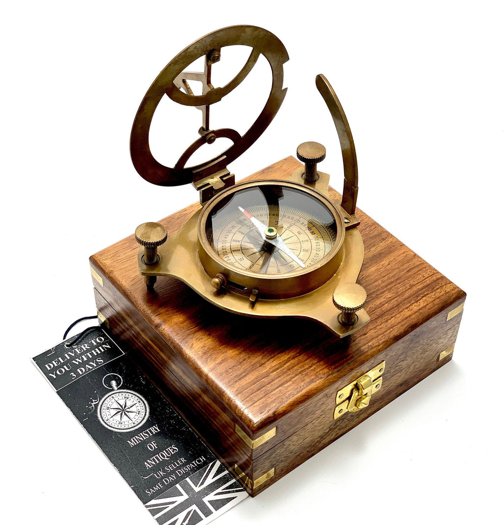 May include: A brass compass and sundial set in a wooden box. The compass features a white face with black markings and a green needle. The sundial has a brass frame with a clear glass cover. The box is made of dark wood with brass hinges and a latch.