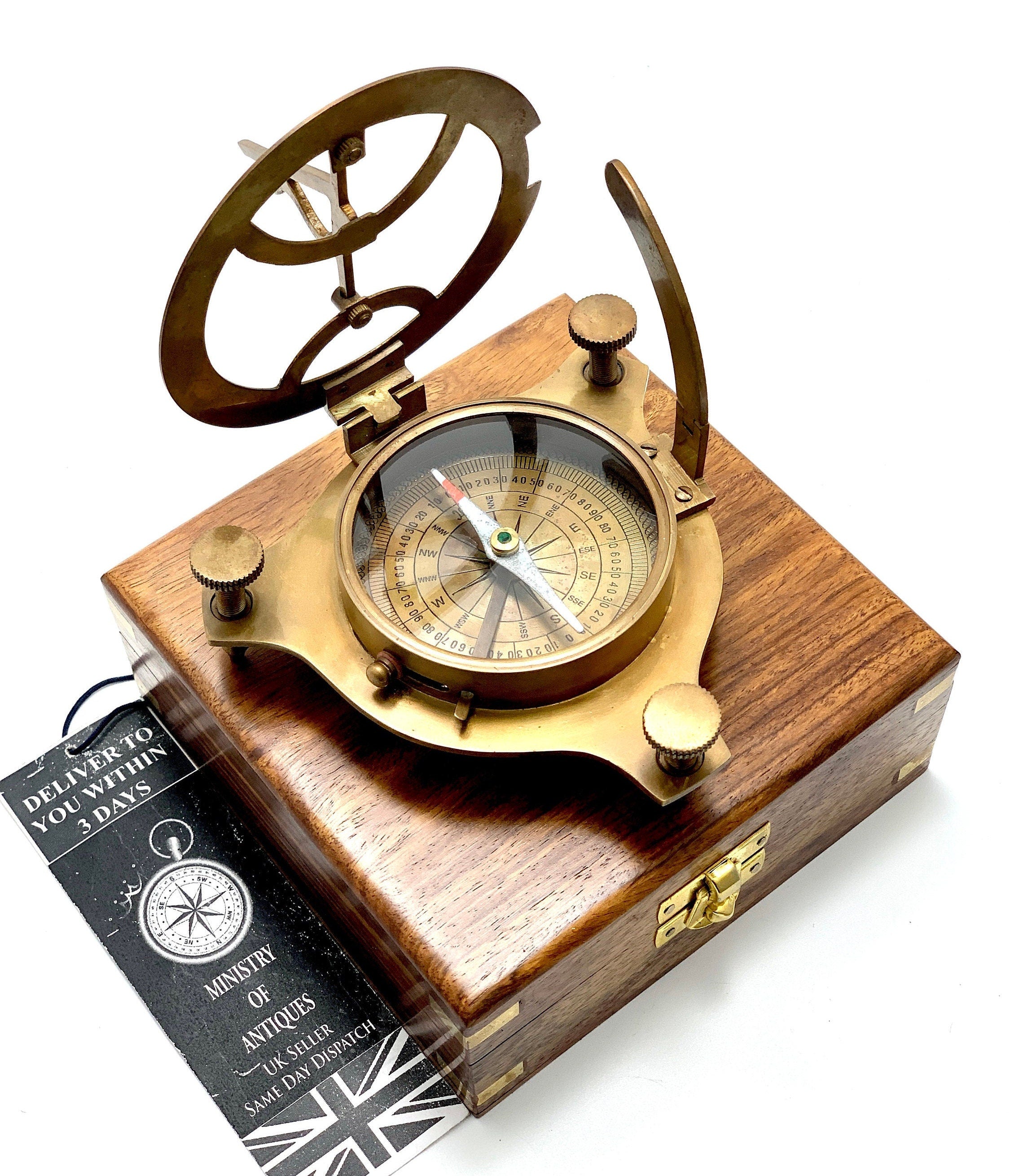 May include: A brass compass and sundial set in a wooden box. The compass features a green needle and a white face with black markings. The sundial has a brass arm and a circular brass face. The box is made of dark wood and has a brass latch.