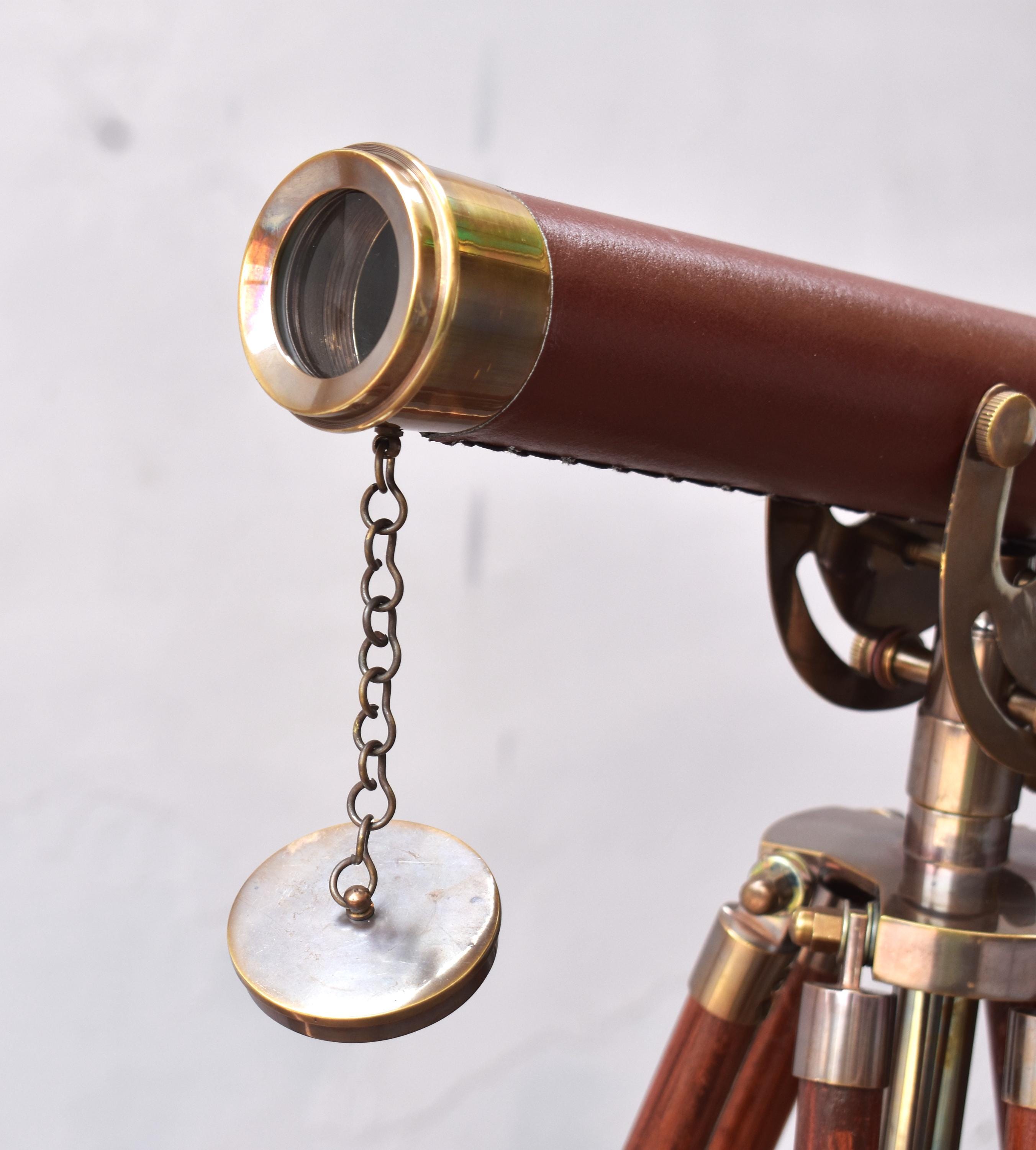 May include: A classic telescope featuring a brown leather barrel and brass detailing. It is mounted on a wooden tripod with brass fixtures. A small, circular lens cap, attached by a chain, hangs below the lens.