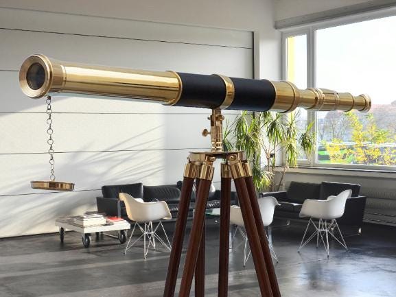 May include: A brass and black telescope on a wooden tripod. The telescope features a gold finish and a black central section. The tripod has dark brown legs with brass detailing. A decorative piece hangs from the telescope.