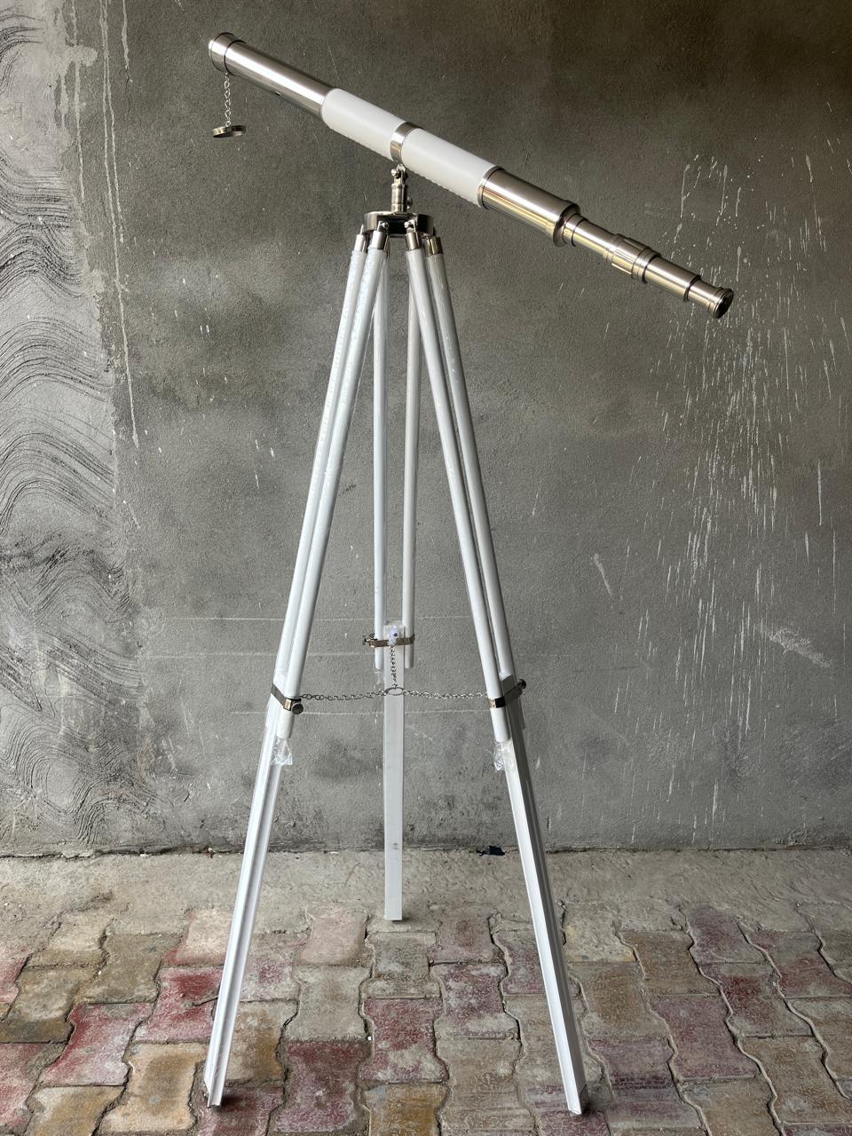 May include: A white and silver telescope on a white tripod. The telescope has a white central section and a silver eyepiece. The tripod has three legs and is connected by a chain. The background is a textured grey wall.