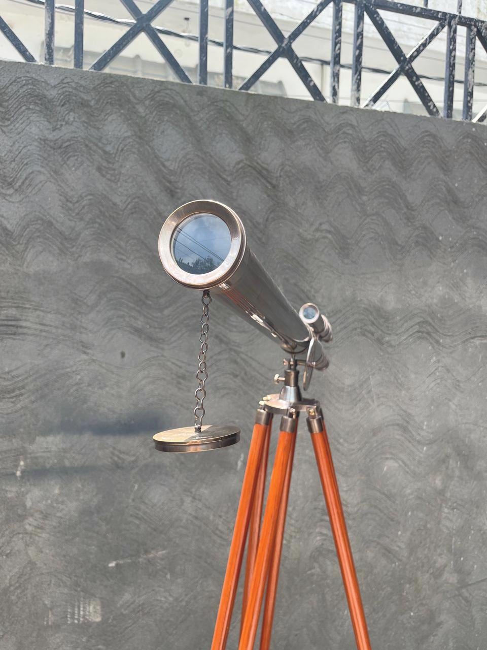 May include: A classic telescope with a silver-coloured body and a small secondary scope. It is mounted on a wooden tripod with orange-brown legs. The telescope has a lens cover attached by a chain.
