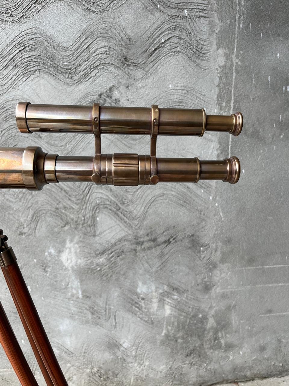 May include: A vintage brass telescope with two parallel barrels, mounted on a wooden tripod. The telescope has a classic design with brass fittings and a weathered appearance. The background is a textured grey wall.