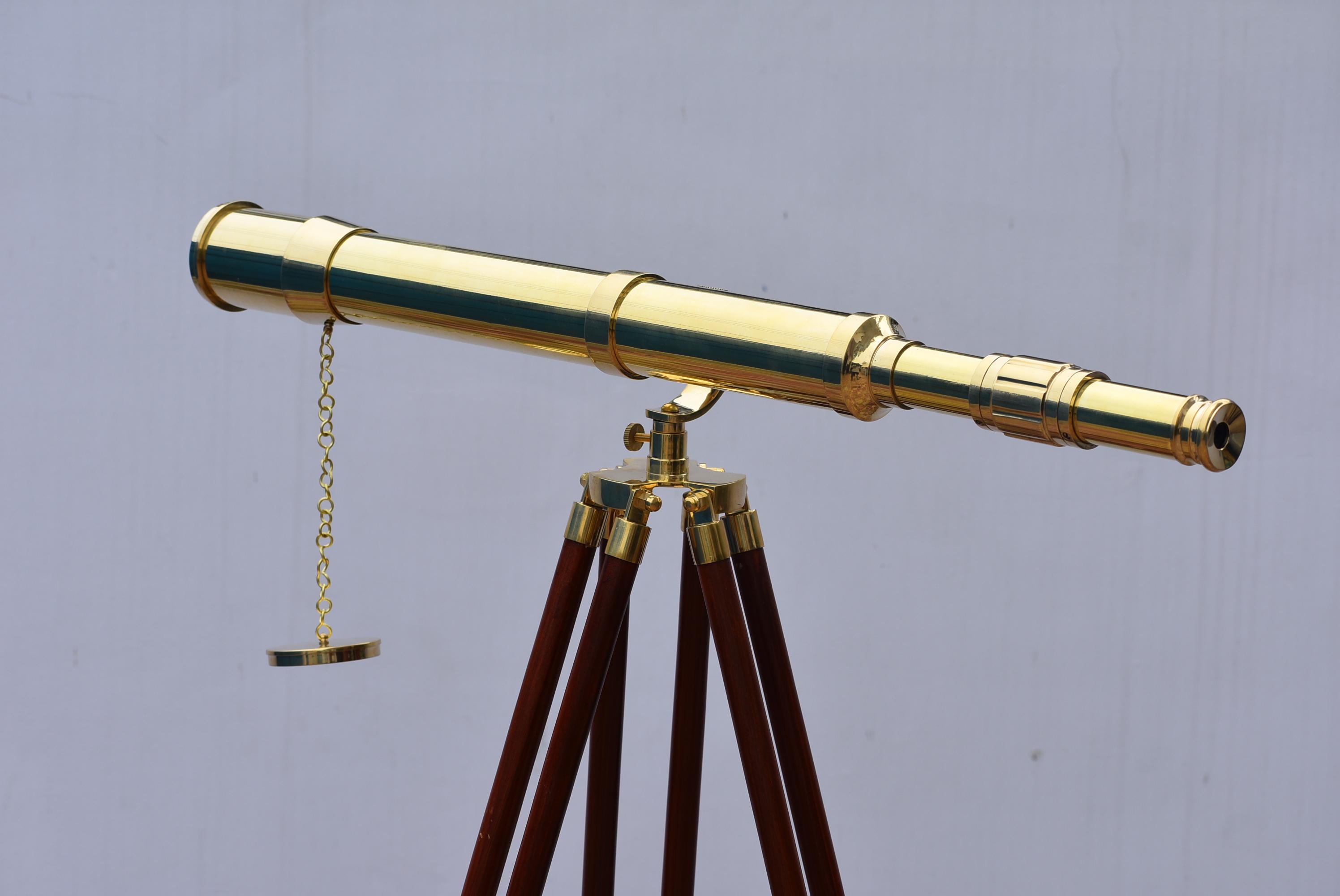 May include: A brass telescope with a gold finish, mounted on a wooden tripod. The telescope has a chain and a small, round brass piece hanging from it. The tripod legs are dark brown.