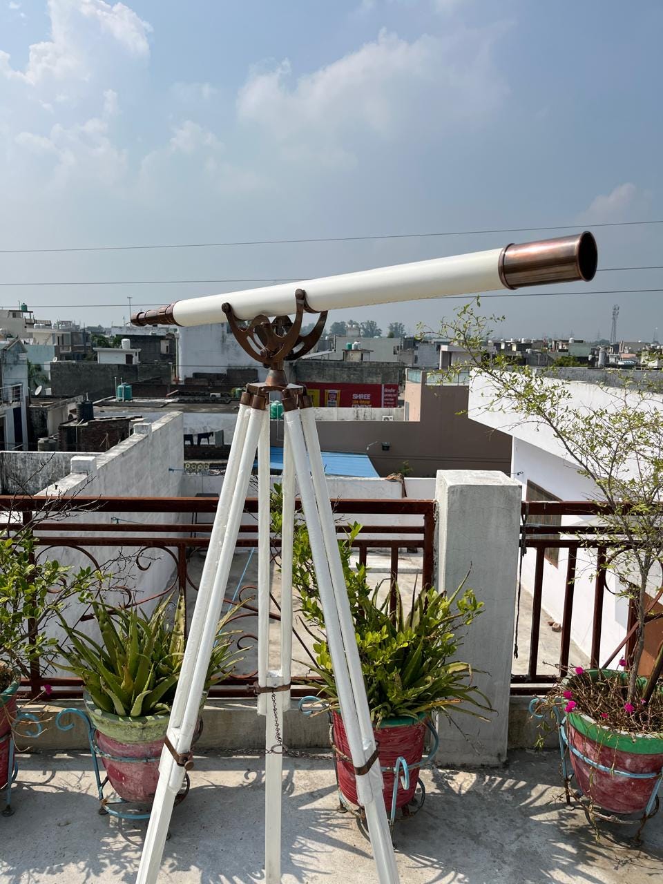 May include: A white and bronze telescope on a white wooden tripod. The telescope features a bronze eyepiece and a white barrel. The tripod has a decorative bronze support. The image is taken outdoors on a sunny day.