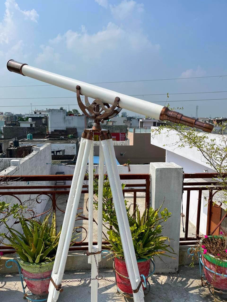 May include: A white and bronze telescope on a white wooden tripod. The telescope features a long, white barrel with bronze detailing and a bronze eyepiece. The tripod has three legs and is linked by a chain.