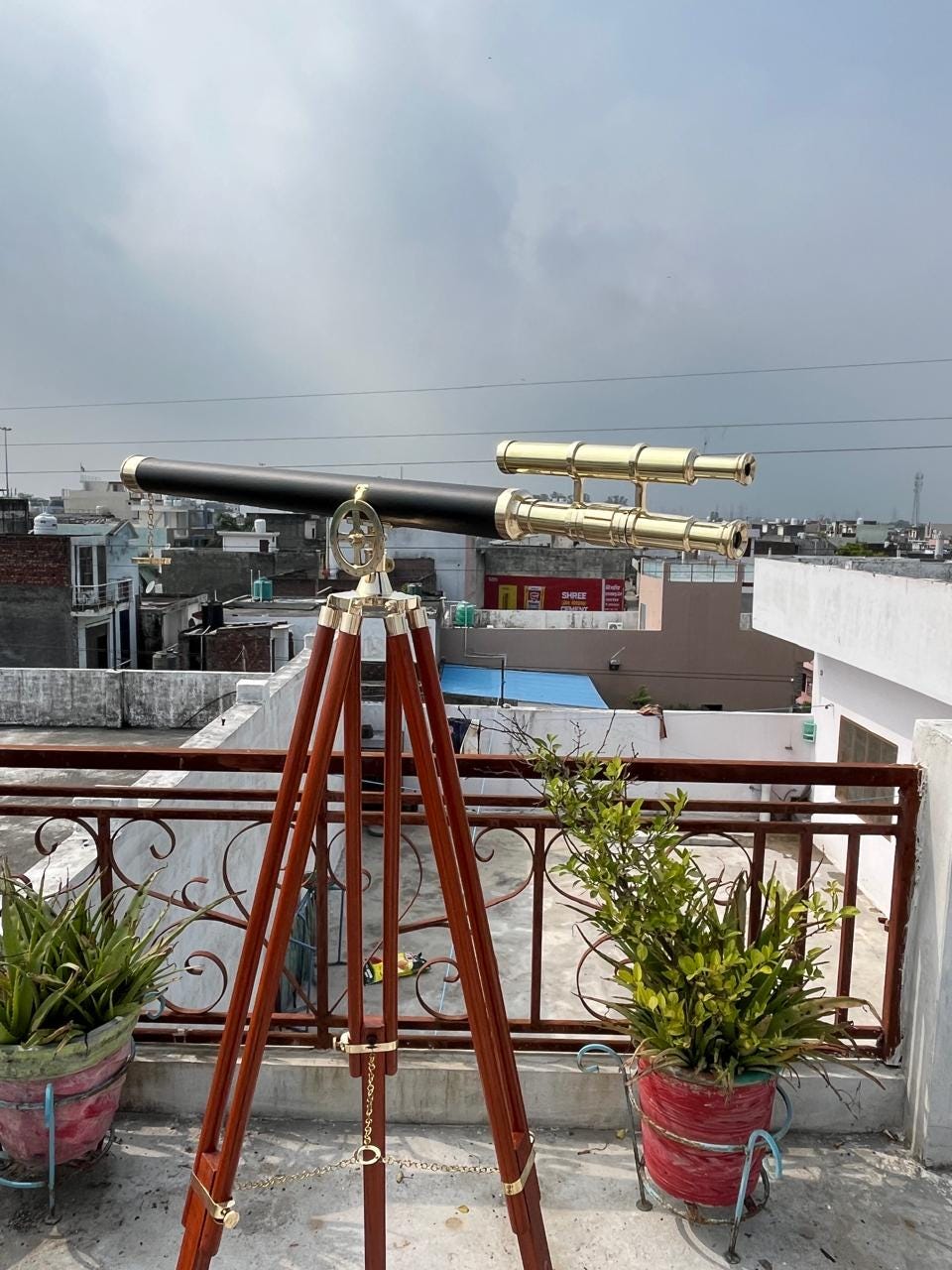 May include: A classic telescope with a black body and brass detailing sits on a wooden tripod. The telescope features a secondary scope. The tripod is made of dark wood and has brass fittings. The scene is set outdoors on a rooftop.