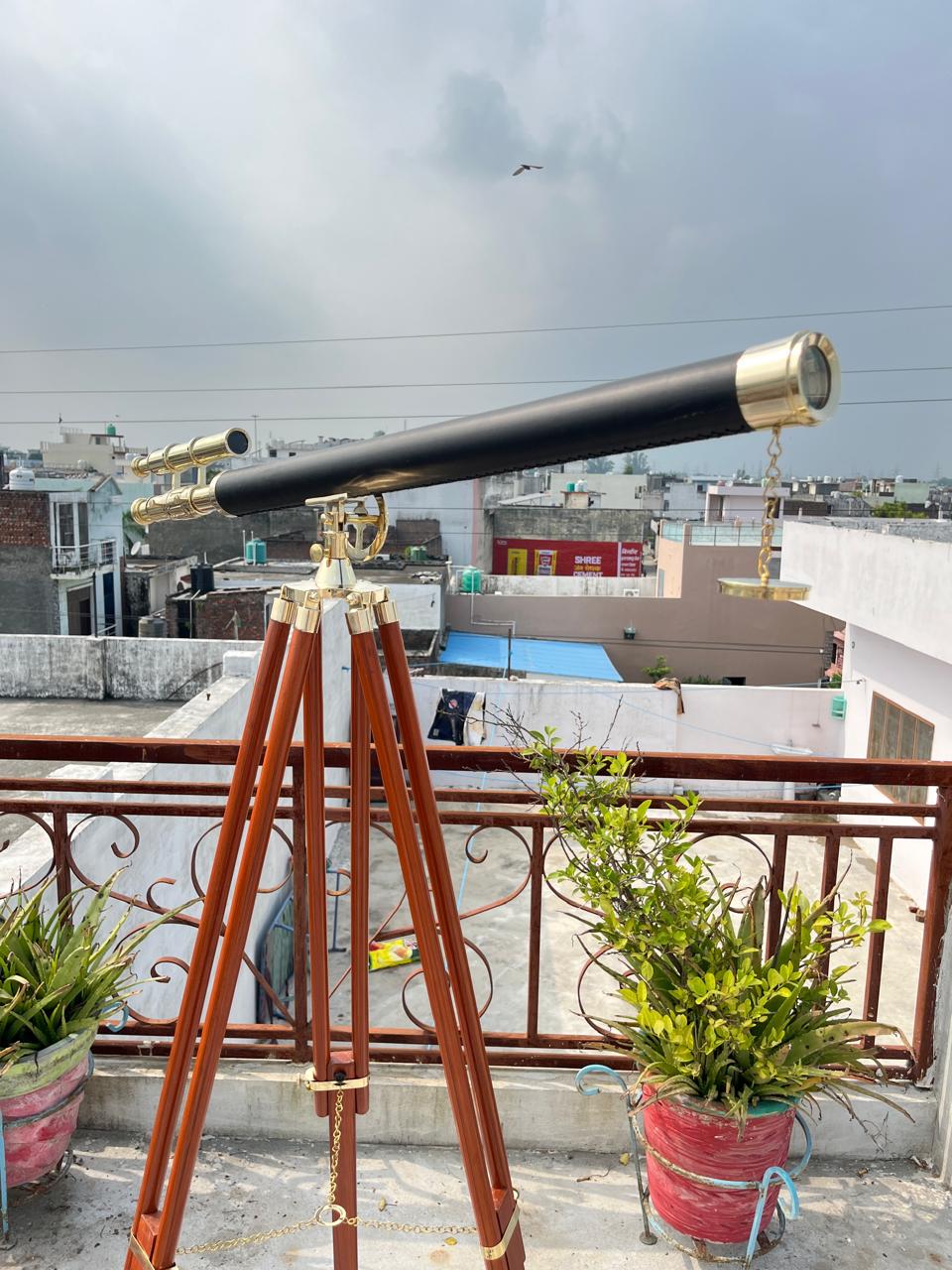 May include: A black and gold telescope on a wooden tripod. The telescope features brass detailing and a black barrel. The tripod is made of dark wood and has brass fittings. The scene is set outdoors with a cloudy sky in the background.