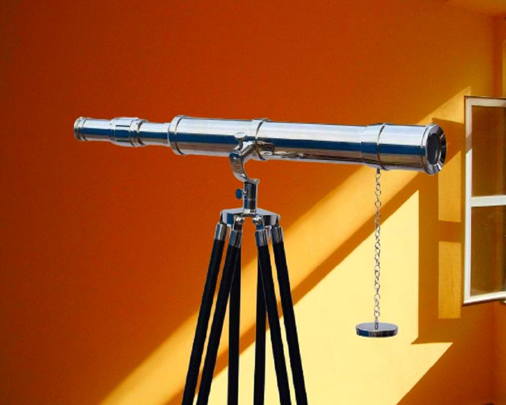 May include: A shiny chrome telescope on a tripod with black legs. The telescope has a chain and a circular weight. The background is an orange wall with a yellow wall and a window.