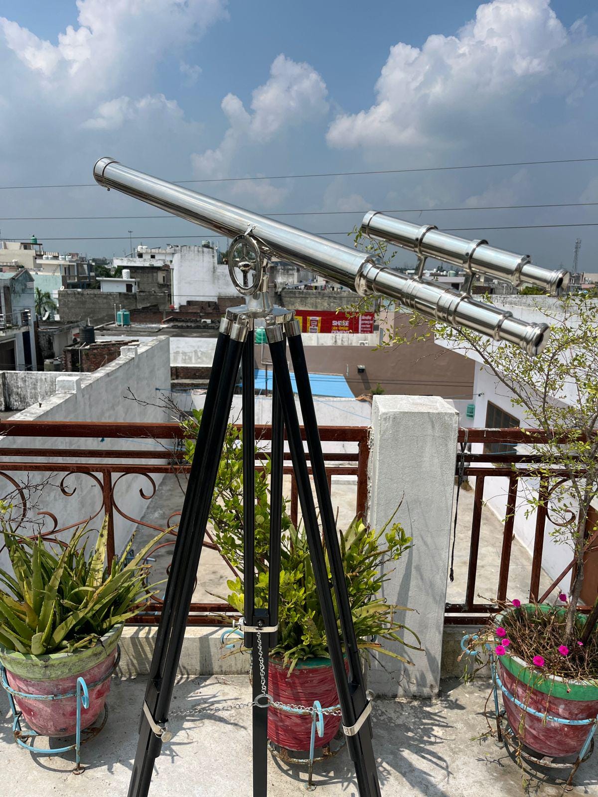 May include: A silver telescope with a black wooden tripod. The telescope has multiple lenses and a detailed design. The tripod has adjustable legs. The background shows a cityscape under a blue sky. The telescope is a classic nautical instrument.