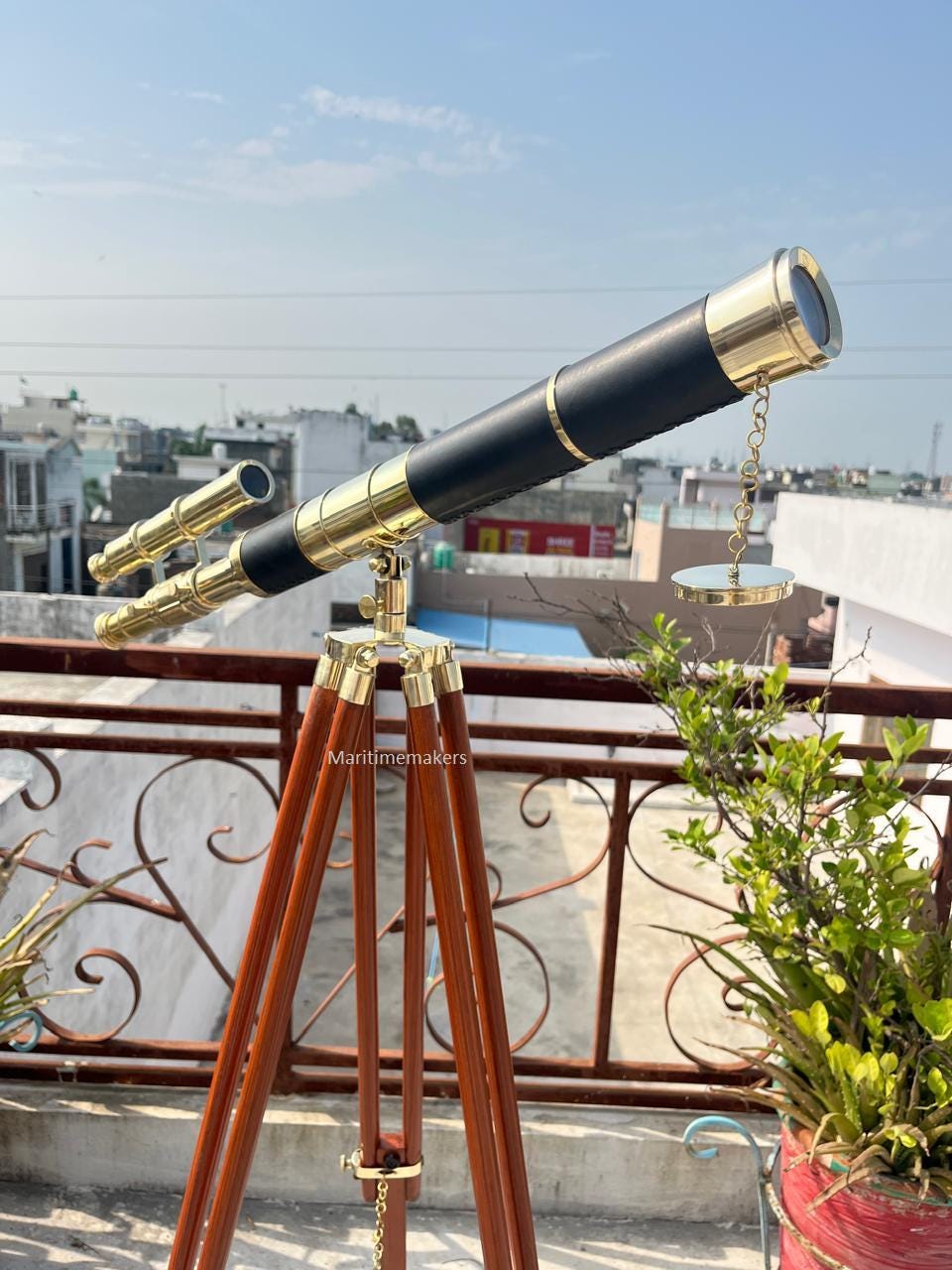 May include: A brass and black telescope on a wooden tripod. The telescope features a long barrel with gold detailing and a smaller secondary scope. The tripod has three brown wooden legs. The text "Maritimemakers" is visible on the tripod.