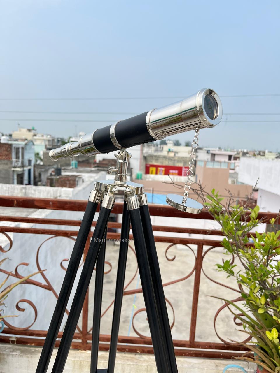 May include: A vintage-style telescope with a black and silver finish, mounted on a wooden tripod. The telescope has a long barrel with a lens at one end and a chain-attached cover. The tripod legs are black and the hardware is silver.