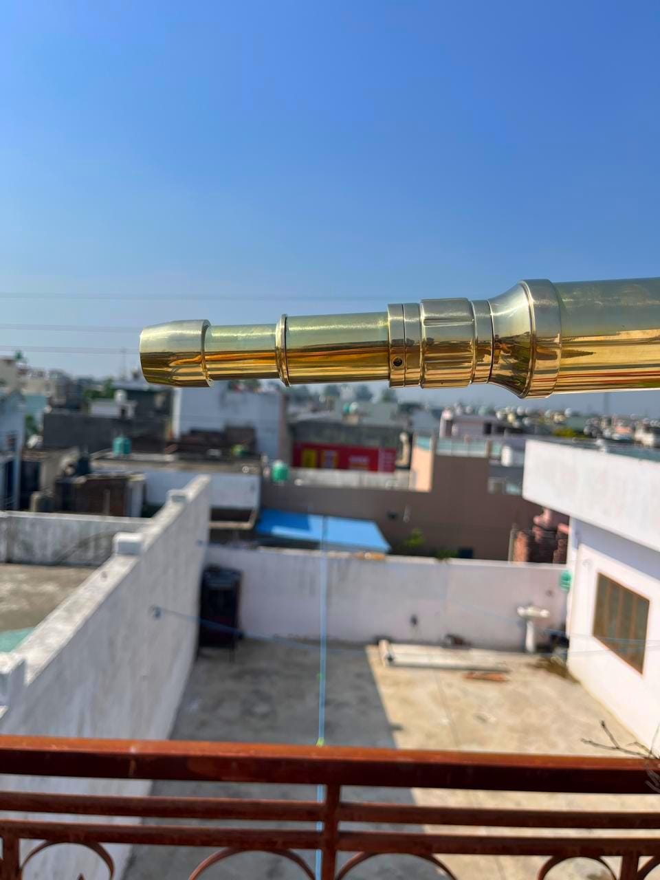 May include: A polished brass telescope, extending towards the left, is set against a clear blue sky. The telescope's design features multiple cylindrical sections and a flared eyepiece. The background shows rooftops and buildings.