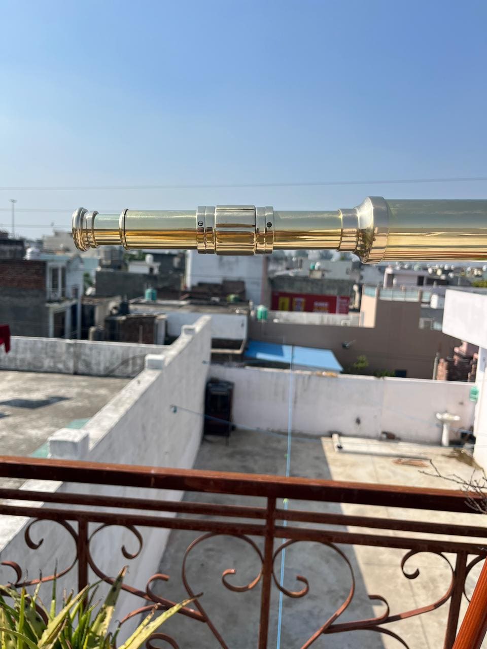 Può includere: Un telescopio in ottone lucido, esteso orizzontalmente, è in primo piano su uno sfondo di tetti e cielo azzurro. La superficie lucida del telescopio riflette la luce solare, evidenziando il suo design e la sua lavorazione.