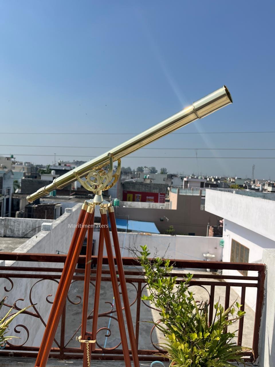 Può includere: Un telescopio in ottone montato su un treppiede in legno. Il telescopio ha una canna lunga e cilindrica inclinata verso l'alto. Il treppiede ha tre gambe in legno scuro. Il cielo è limpido e blu.