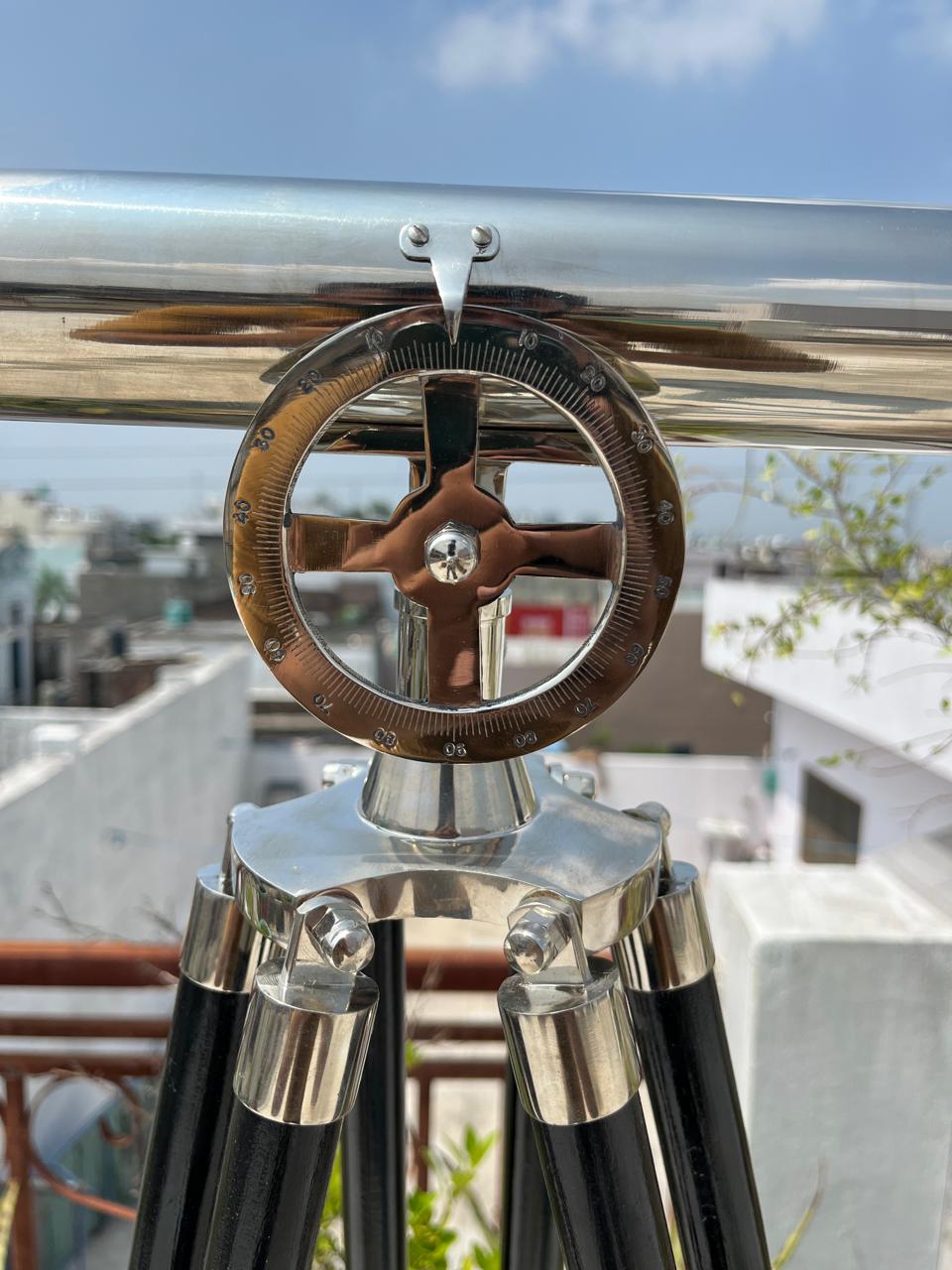 May include: A silver and black telescope mounted on a tripod. The telescope features a brass dial with degree markings and a cross-shaped handle. The tripod legs are black with silver detailing. The background shows a sunny outdoor scene.