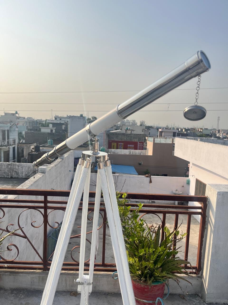 Può includere: Un telescopio argentato e bianco montato su un treppiede bianco. Il telescopio ha una canna lunga e cilindrica con una lente a un'estremità e un piccolo coperchio rotondo appeso a una catena. Il treppiede ha tre gambe.