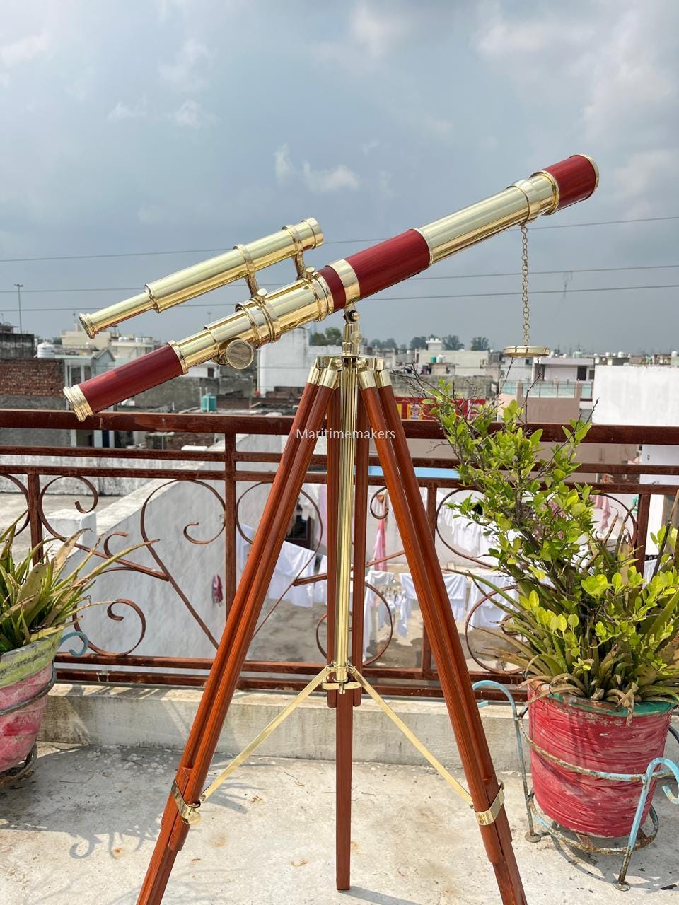 May include: A brass and wood telescope with a red barrel and a smaller brass scope attached. The telescope is mounted on a wooden tripod with brass accents. The image is taken outdoors on a sunny day.