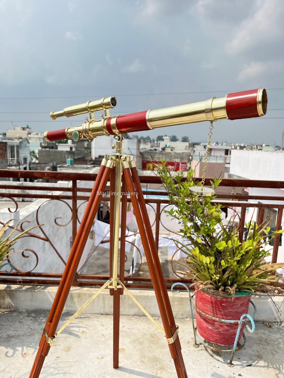May include: A brass and red telescope mounted on a wooden tripod. The telescope has a long barrel with a red section and a smaller, angled eyepiece. The tripod has brass accents and is set up outdoors.