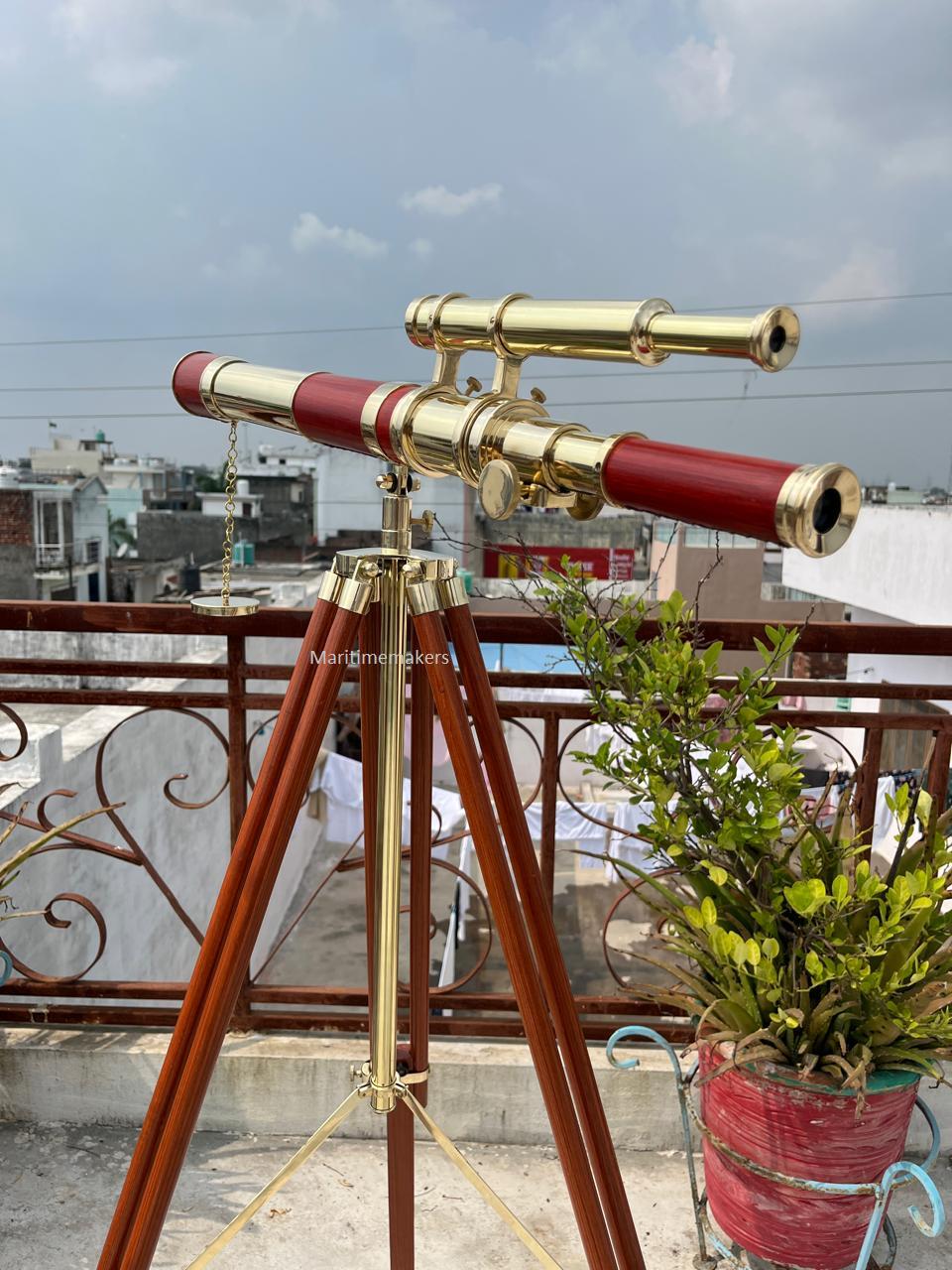 May include: A brass and red telescope mounted on a wooden tripod. The telescope has a gold finish with red accents and is positioned outdoors. The tripod has dark brown legs and brass hardware. The background shows a cityscape and a cloudy sky.