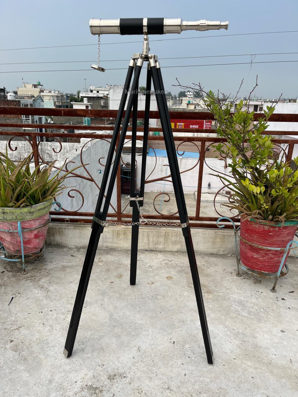 May include: A vintage-style telescope with a silver and black finish, mounted on a black wooden tripod. The telescope has a classic design with a long barrel and adjustable focus. The tripod has metal accents and a chain for stability. The setup is outdoors.