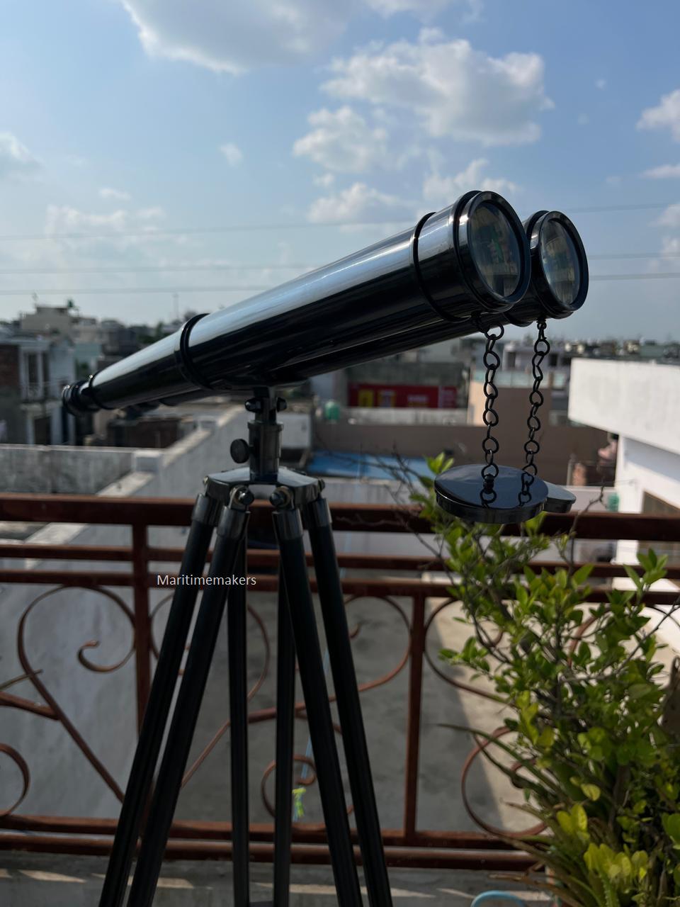 Può includere: Un telescopio nero con finitura metallica lucida, montato su un treppiede nero. Il telescopio ha due oculari ed è collegato a un piccolo coperchio rotondo tramite catene. Il treppiede ha tre gambe. Il testo "Maritimemakers" è visibile.
