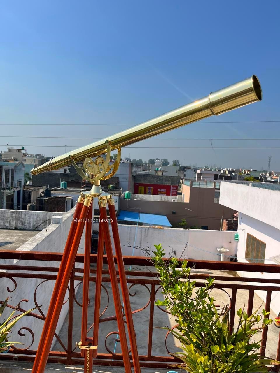 May include: A brass telescope mounted on a wooden tripod. The telescope has a long, cylindrical barrel and is supported by a decorative brass mount. The tripod legs are a dark reddish-brown color. The sky is a clear blue.