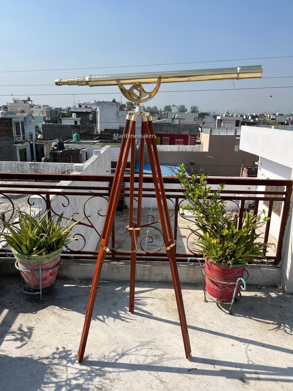 May include: A brass telescope mounted on a wooden tripod. The telescope has a long, cylindrical barrel and is supported by a decorative brass mount. The tripod legs are dark brown and adjustable. The image is taken outdoors on a sunny day.