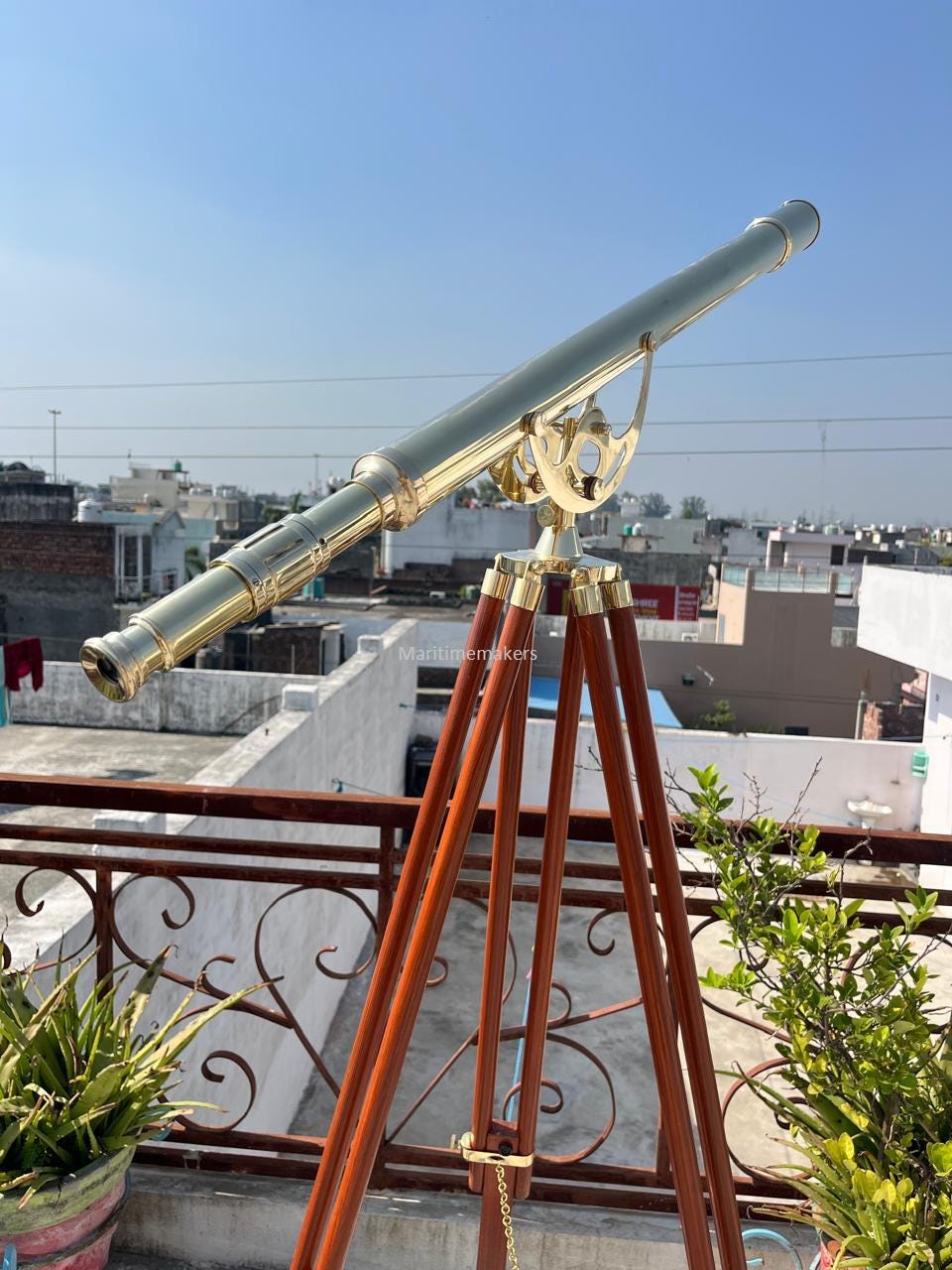 Può includere: Un telescopio in ottone montato su un treppiede in legno. Il telescopio ha una canna lunga e cilindrica inclinata verso l'alto. Le gambe del treppiede sono di colore marrone scuro. Lo sfondo mostra un cielo blu chiaro e tetti.