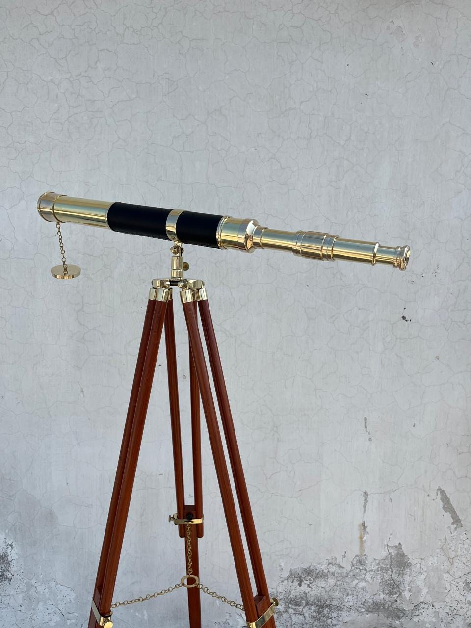 May include: A brass telescope with a black leather grip is mounted on a wooden tripod. The telescope has a gold finish and a chain with a small circular piece attached. The tripod has dark brown legs and gold accents.