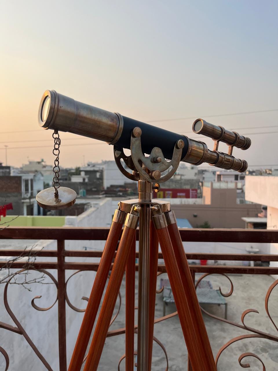 Può includere: Un telescopio in ottone in stile vintage con un corpo nero, montato su un treppiede in legno. Il telescopio ha un design classico con una catena e un piccolo coperchio rotondo in ottone. Le gambe in legno sono di colore marrone scuro e gli accenti metallici sono dorati.