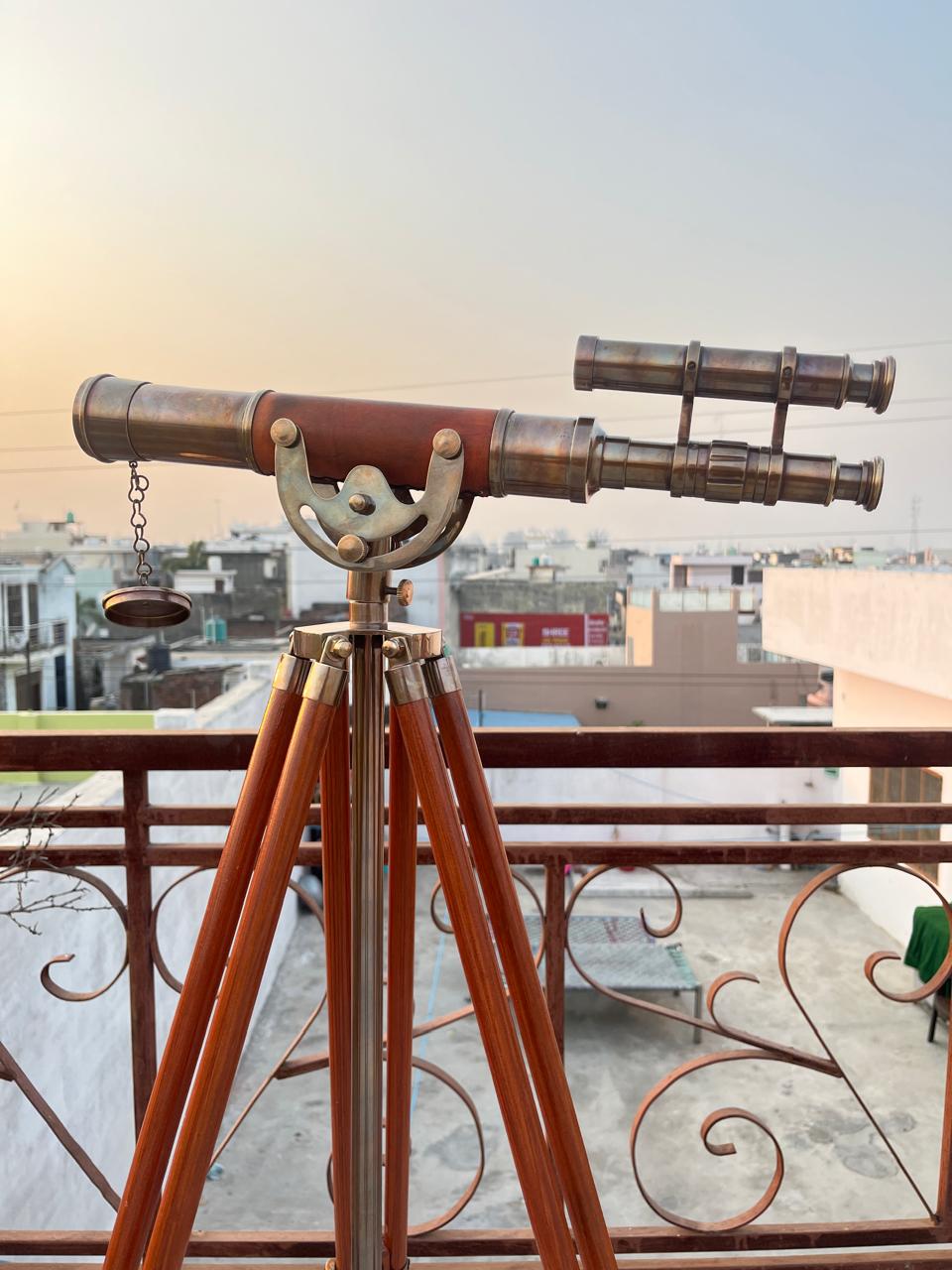 May include: A classic brass telescope mounted on a wooden tripod. The telescope features a dark brown wooden barrel and brass fittings. The tripod legs are made of polished wood. The background shows rooftops and a clear sky.