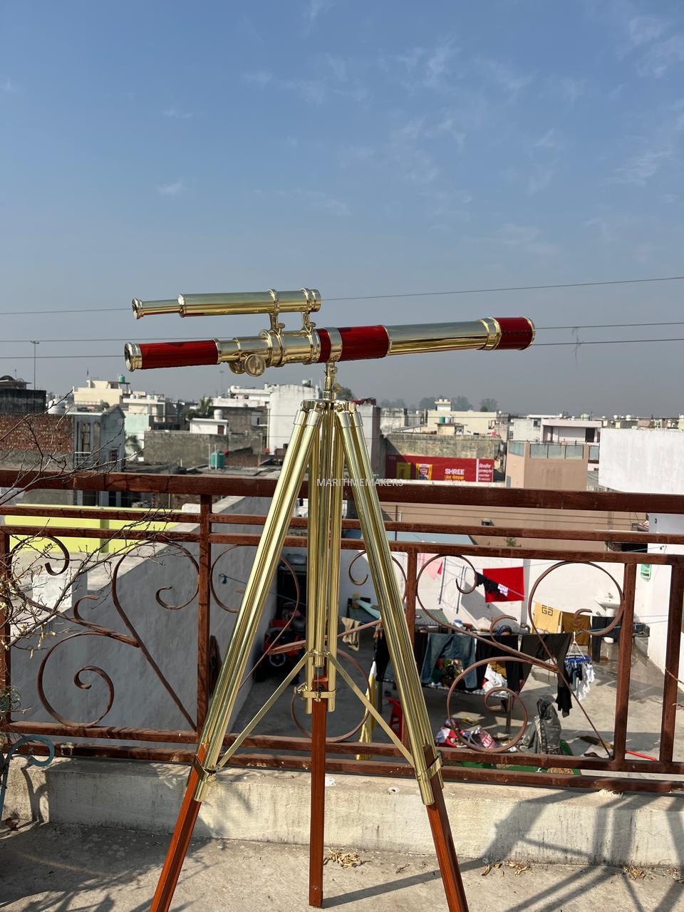 May include: A brass and red telescope on a wooden tripod. The telescope features a long barrel with red detailing and a smaller, angled eyepiece. The tripod has brass legs and wooden supports, set against a clear blue sky.