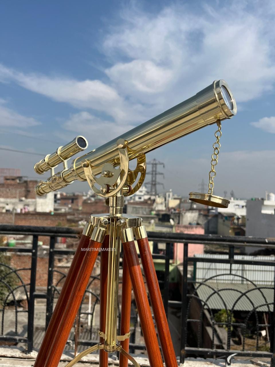 May include: A brass telescope mounted on a wooden tripod. The telescope has a long, cylindrical barrel with a smaller eyepiece and a chain-connected lens cap. The tripod legs are a rich brown color, and the sky is blue.