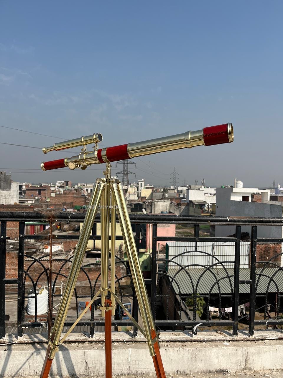 May include: A brass and red telescope on a wooden tripod. The telescope features a long barrel with red detailing and a smaller, parallel scope. The tripod has brass legs and wooden supports, set against a cityscape and a blue sky.