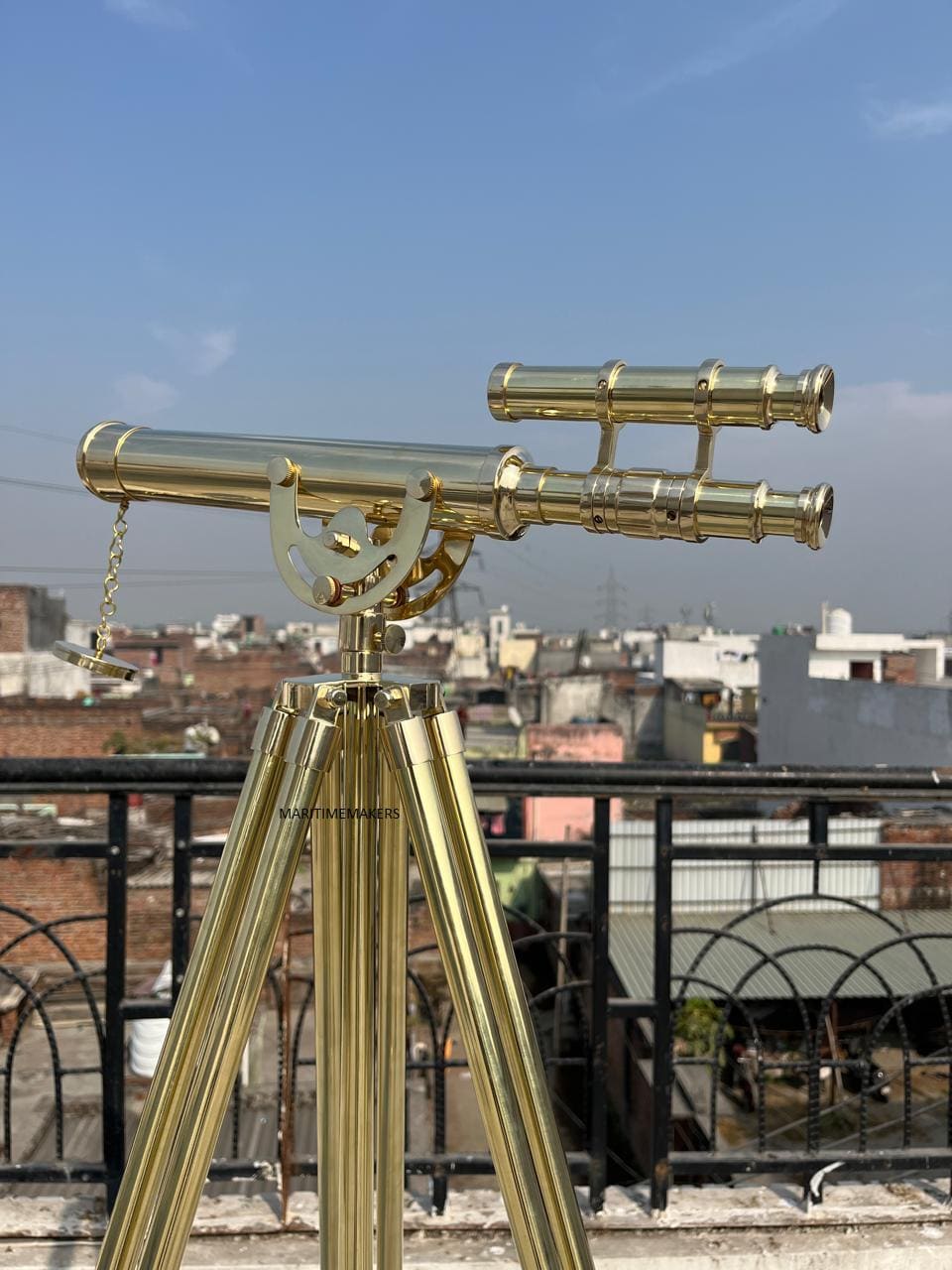 May include: A brass telescope mounted on a tripod, set against a cityscape and blue sky. The telescope has a long barrel with a chain and a smaller, parallel scope. The tripod has three legs.