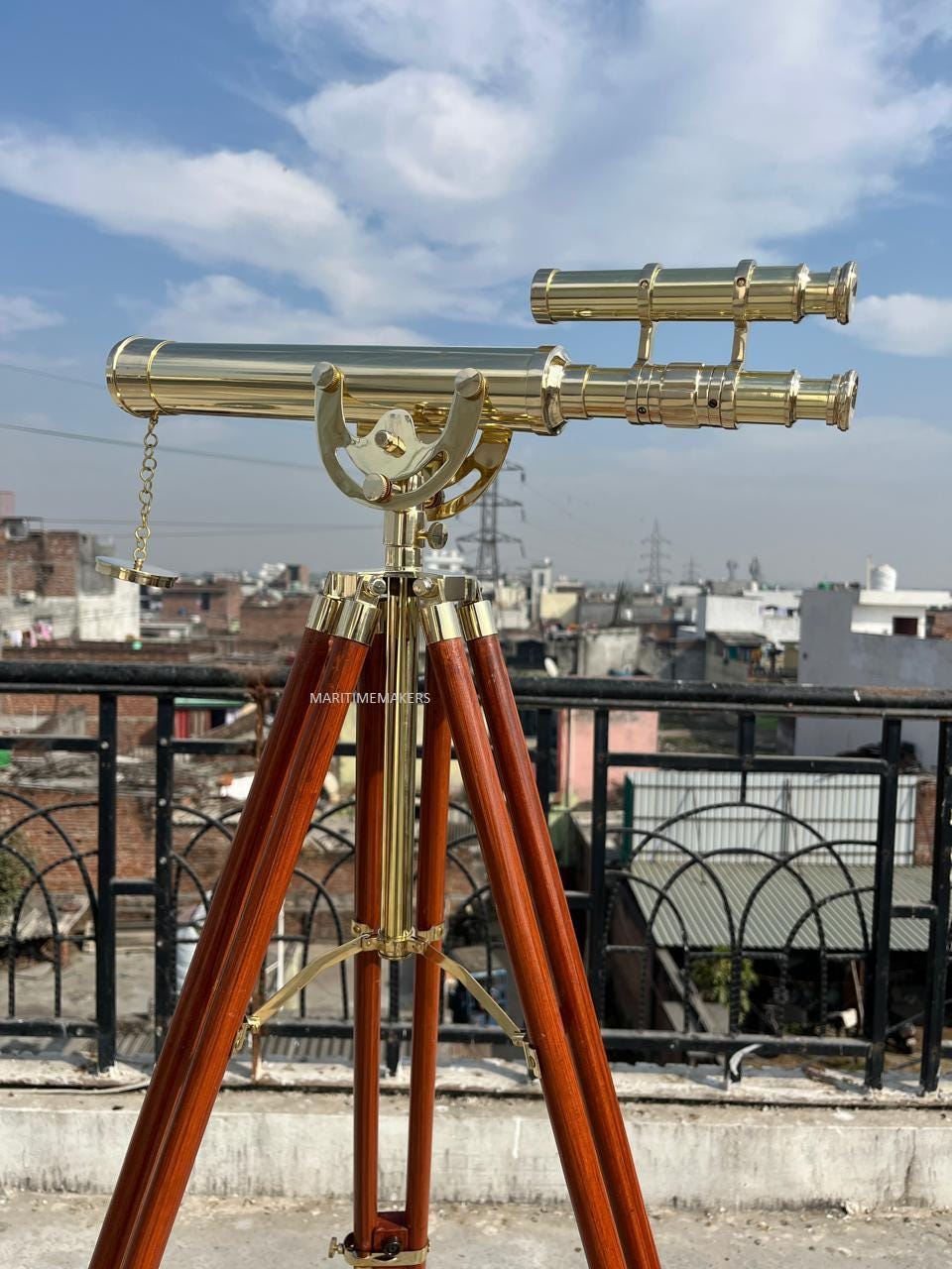 May include: A brass telescope mounted on a wooden tripod. The telescope has a long barrel and a smaller, parallel scope. The tripod legs are a dark brown color. The text "MARITIMEMAKERS" is visible on the tripod.